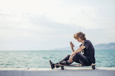 telefonunu kullanarak longboard üzerinde oturan hipster adam