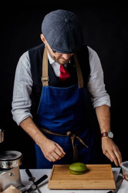 Aşçı stüdyoda, siyah duvara karşı tahta bir masanın yanında duruyor..