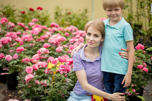 Beautiful young woman with short blond hair posing with son near roses in garden