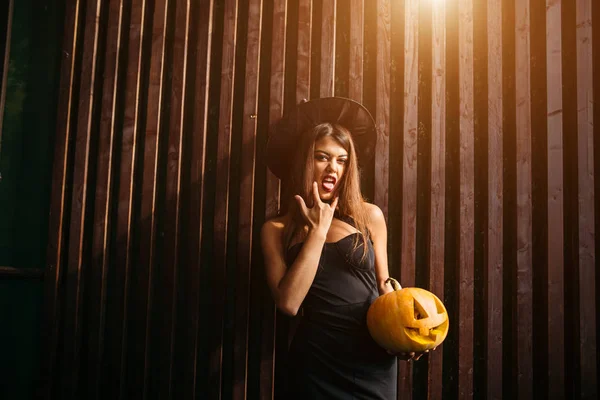 Halloween Witch with a carved Pumpkin. Beautiful young woman in witches hat and costume holding pumpkin.
