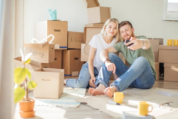 Coppia che si trasferisce in una nuova casa. Seduto sul pavimento e rilassante dopo lo spacchetto.Fare selfie con smartphone — Foto Stock