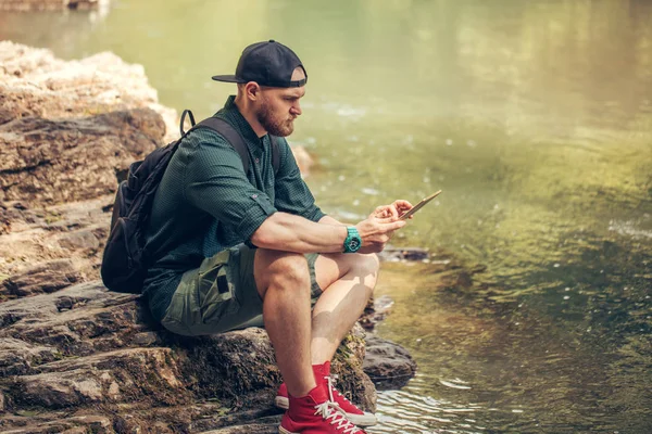 Jeden muž turista pomocí tabletu na přírodě, zatímco sedí na skalnatém břehu řeky — Stock fotografie