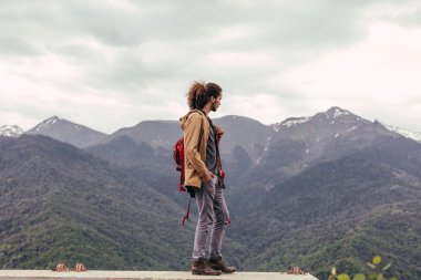 Kırmızı sırt çantalı gezgin dışarıda dinleniyor ve arka planda kayalık dağlar var.