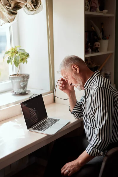 Dojrzały mężczyzna wygląda wyczerpany siedząc przy laptopie i trzymając okulary w dłoni — Zdjęcie stockowe