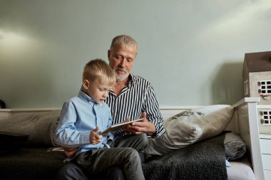 Grandfather and grandson using digital tablet while sitting on couch clipart