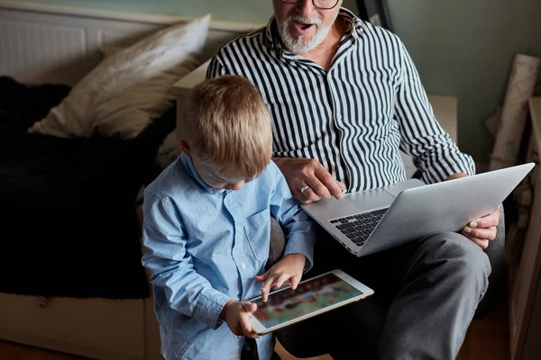 Vovô e neto usando gadgets e sorrindo enquanto passam o tempo juntos sentados no sofá — Fotografia de Stock