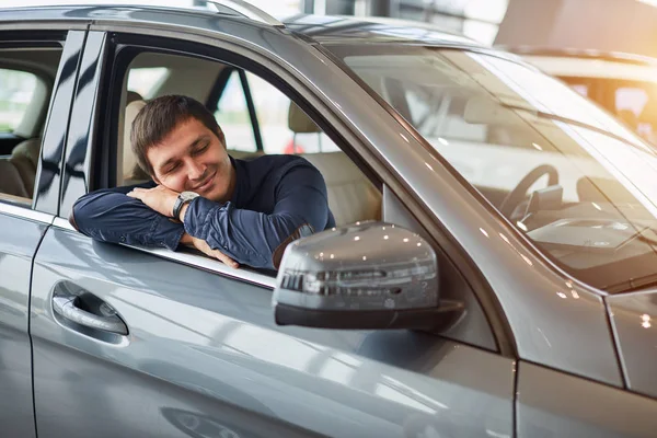 Посещение автосалона. Мечтающий мужчина обнимает новую машину и улыбается — стоковое фото