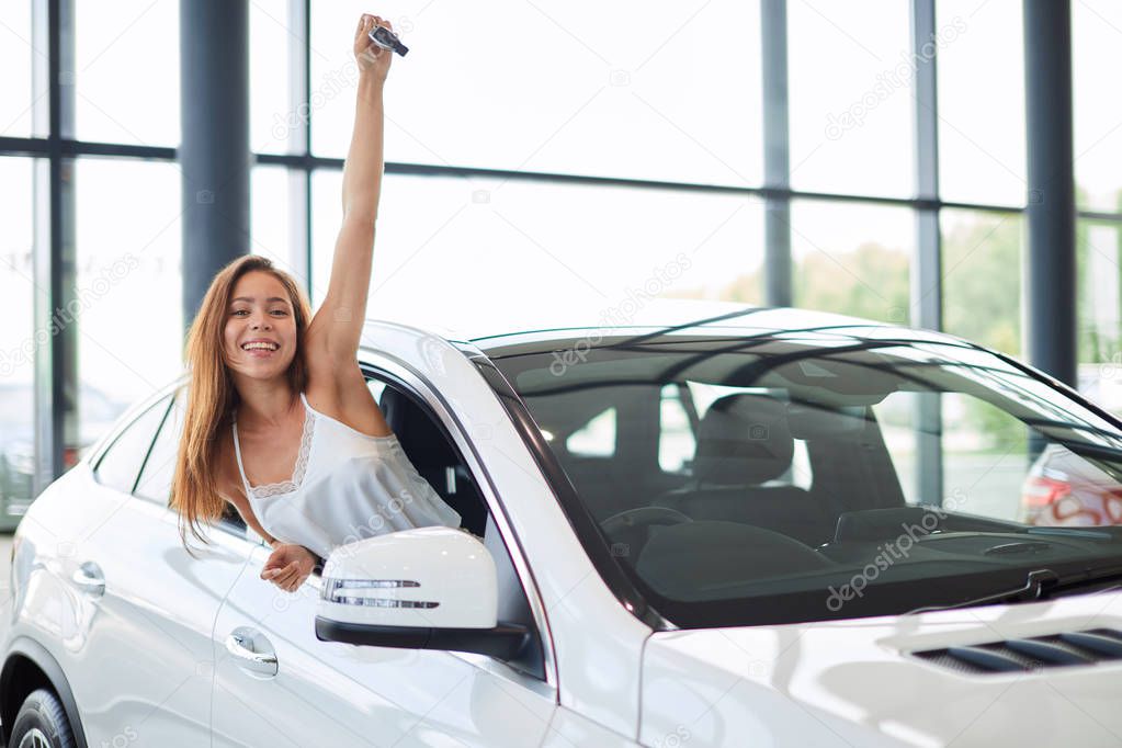 Happy woman driver in white car holds car keys in her new car at car show room