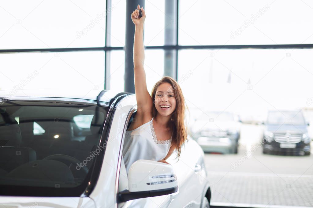 Happy woman driver in white car holds car keys in her new car at car show room