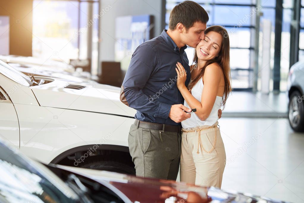 Happy loving couple embracing near their new luxury car at dealership.