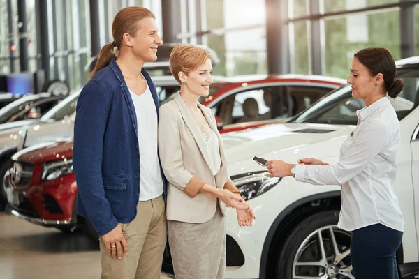 Feliz joven pareja recibiendo la llave del coche de un vendedor en el concesionario de automóviles . — Foto de Stock