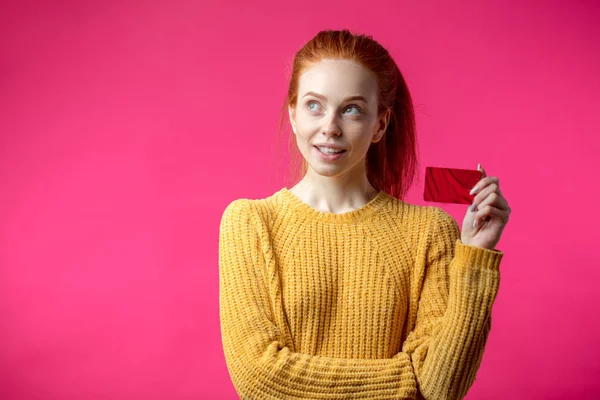 Feliz hermosa mujer de jengibre sosteniendo tarjeta de crédito aislada sobre fondo rosa . —  Fotos de Stock