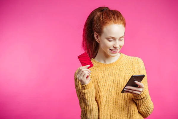 Портрет милой рыжей девушки с помощью смартфона на розовом фоне — стоковое фото