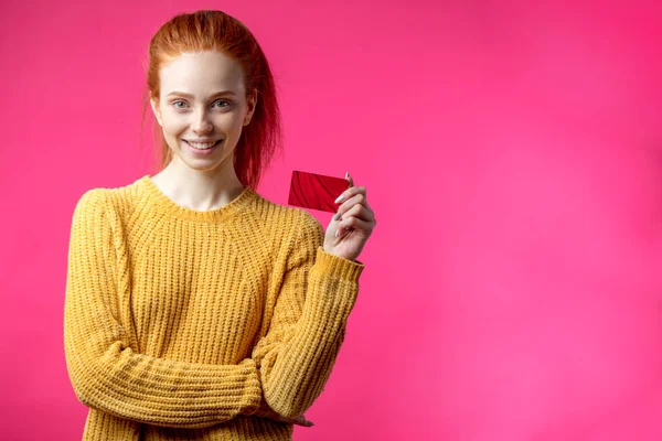 Feliz hermosa mujer de jengibre sosteniendo tarjeta de crédito aislada sobre fondo rosa . —  Fotos de Stock