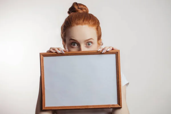 Portrait of cheerful redhead girl with hair bun holding empty wooden frame. — Stock Photo, Image