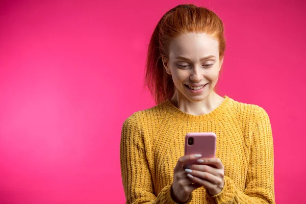 Портрет милой рыжей девушки с помощью смартфона, изолированного на розовый б — стоковое фото