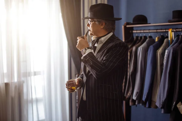 上流階級のシックな精悍な億万長者や喫煙タバコ パイプ Buing 新しい排他的なスーツ 高級ファッション ブティックを訪問豊かな成熟した男の肖像画 準備ができたと服のラックを作った背景にスーツをマン — ストック写真