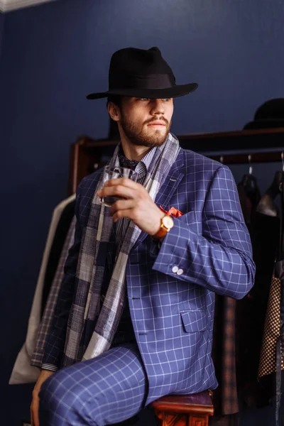Homem elegante em terno de moda e chapéu posando no atelier com bebida — Fotografia de Stock