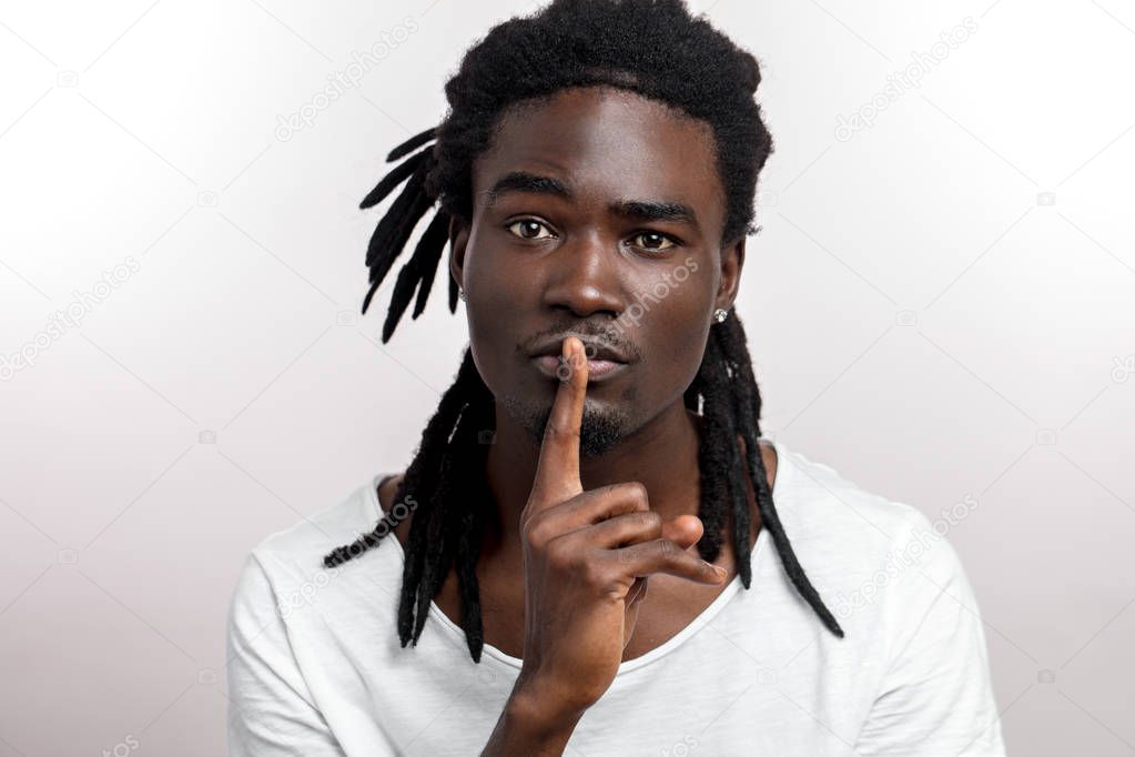dark-skinned young man gesturing as secret holding his index finger at his lips, saying Shh