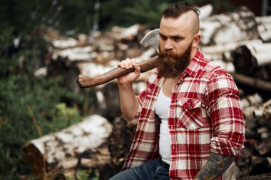 Sakallı mohawk adam yakacak odunluğunun yanında duruyor.