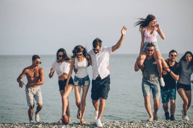 Bir grup arkadaş sahilde yürüyor, eğleniyor, kadınlar erkekleri sırtında taşıyor, komik bir tatil.