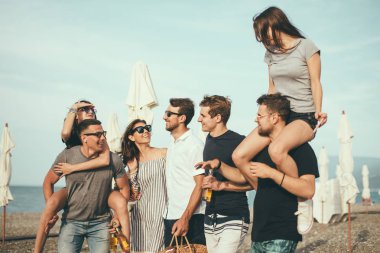 Bir grup arkadaş sahilde yürüyor, eğleniyor, kadınlar erkekleri sırtında taşıyor, komik bir tatil.