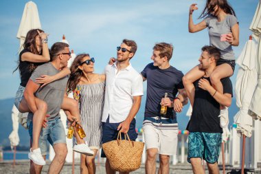 Bir grup arkadaş sahilde yürüyor, eğleniyor, kadınlar erkekleri sırtında taşıyor, komik bir tatil.