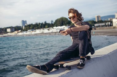 telefonunu kullanarak longboard üzerinde oturan hipster adam