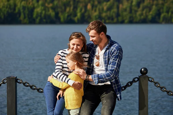 Щаслива сім'я проводить час разом біля озера. Батьки грають з дочкою обіймаються і розважаються — стокове фото