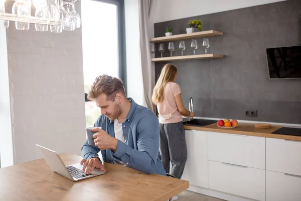 Lachende jonge man aan het werk op laptop en zijn vriendin de afwas — Stockfoto