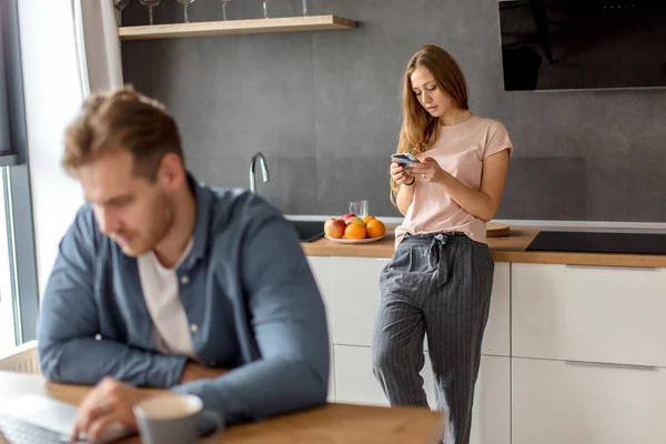 Ehrfürchtige blondhaarige Hausfrau schickt SMS, während ihr Freund das Notizbuch benutzt — Stockfoto