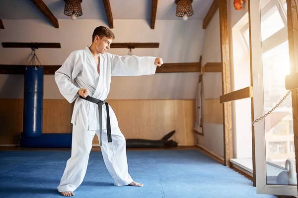 Hombre en kimono blanco con cinturón negro entrenamiento karate en gimnasio — Foto de Stock
