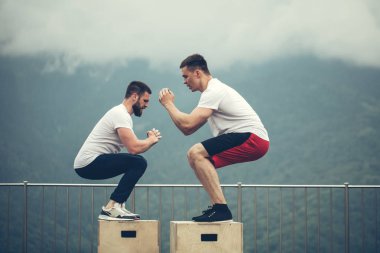 Genç erkek eğitmen ve sportif öğrencisi kutusunda egzersiz rutin bir parçası olarak atlama beyaz haç uygun yerleştirmek. Taze dağ havası üzerinde açık egzersiz atlama kutu. Etkin ve sağlık yaşam kavramı