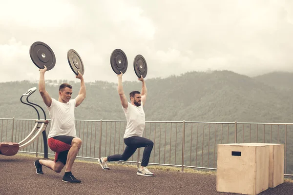 Two athletes with barbell disc making lunges in outdoor workout — Stock Photo, Image