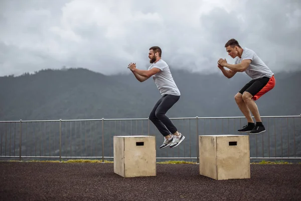 남자 운동 친구 두 명이 산 위 야외에서 박스 점프를 하는 모습. — 스톡 사진