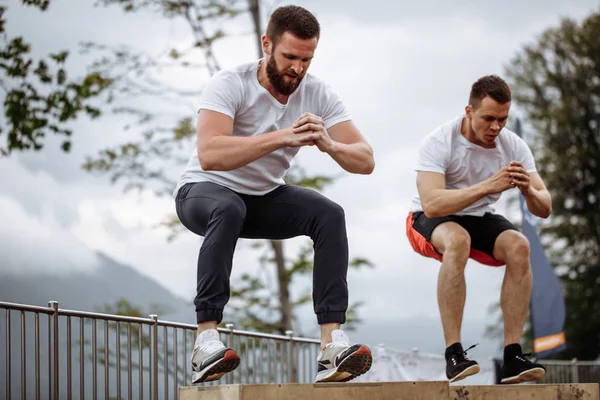 Två manliga atletiska vänner gör box hoppa utomhus på toppen av berget. — Stockfoto
