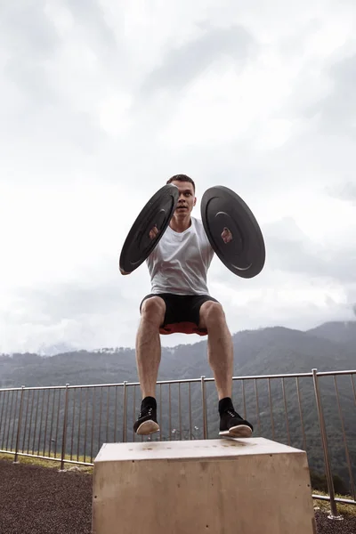 Strong male athlete doing box jumps with two plates outdoor on top of mountain. — Stock Photo, Image