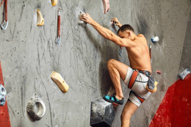 Male climber exercising in gym, climbing up on artificial wall with boulders. clipart