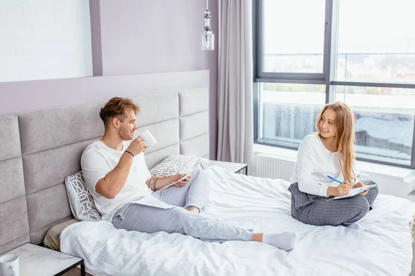 Schöner lächelnder Mann plaudert mit seiner attraktiven Freundin, während er auf dem Bett sitzt — Stockfoto