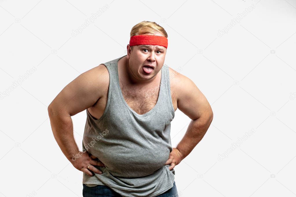 Obese unsporting non- athletic man during his training session, resting, exhausted, taking a overwork, wipes sweat with hand from his forehead against white background. I