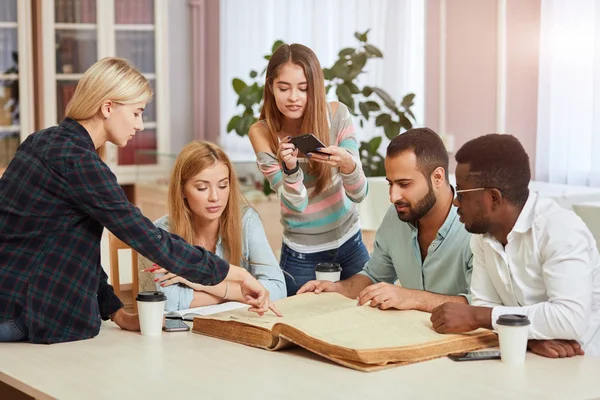 Diversos estudiantes multirraciales pasar el tiempo libre en la biblioteca con gran libro viejo — Foto de Stock