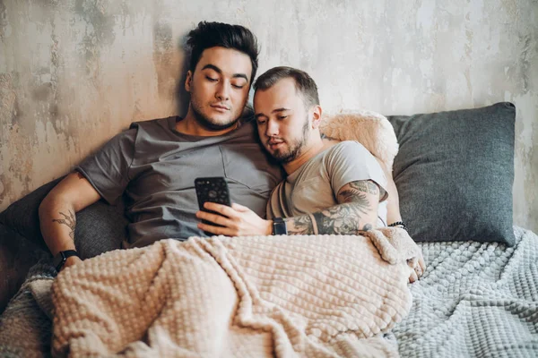 Felice coppia gay sdraiato insieme a letto e utilizzando smartphone, guardando le foto — Foto Stock