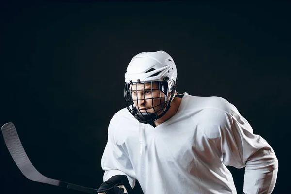 Isolated shot of ice-hockey player in white sportswear and white helmet holding hockey stick prepare to defense on black background