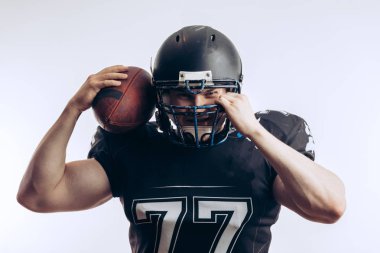 Koruyucu üniformalı ve kasklı Amerikan futbolcusu elinde top tutuyor.