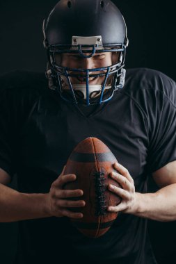 Siyah arka planda topu olan siyah spor giyimli Amerikalı bir futbolcu.