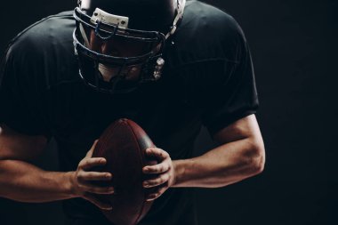 Kasklı ve zırhlı Amerikan futbolcu siyah duvara karşı top taşıyor.