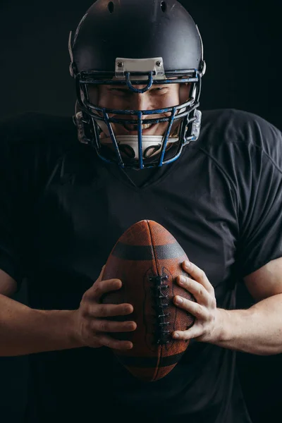 American football player in black sportwear with a ball on black background