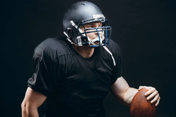 Jugador de fútbol americano en ropa deportiva negra con una pelota sobre fondo negro — Foto de Stock