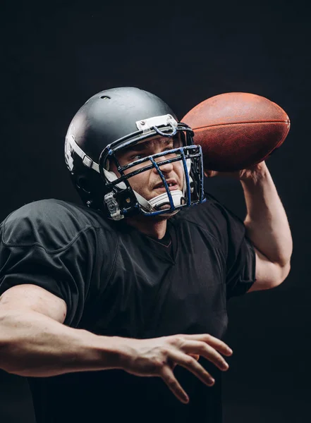 Jugador de fútbol americano con casco y armadura con una pelota contra la pared negra — Foto de Stock