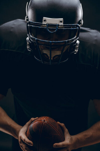 American football player with helmet and armour with a ball against black wall